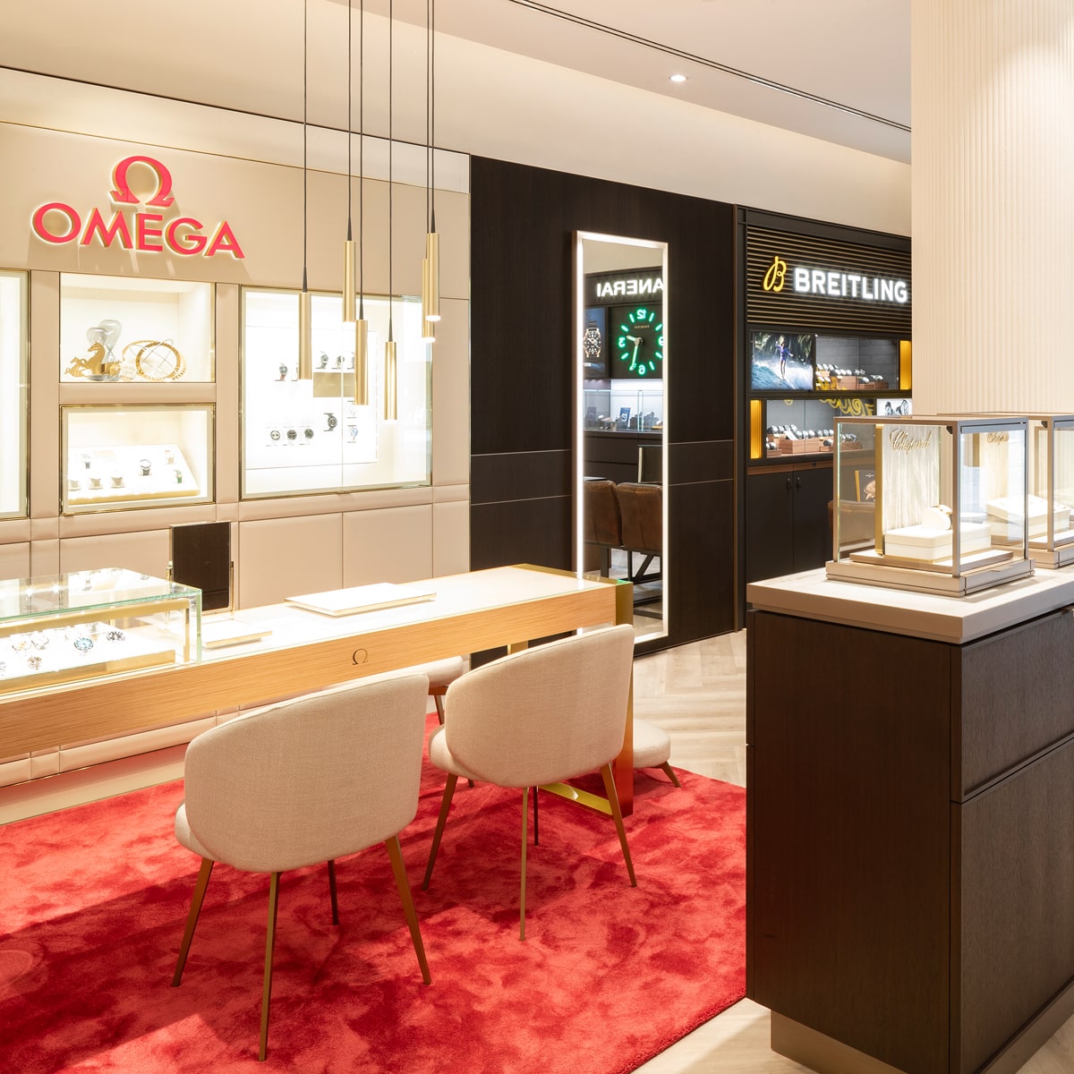 © Juwelier Wenthe Luxusuhrenboutique mit Uhrenmarken wie Omega und Breitling, zwei beigefarbenen Stühlen an einem Holztisch auf einem roten Teppich und hell beleuchteten Glasvitrinen, in denen Uhren präsentiert werden. Modernes und elegantes Design.