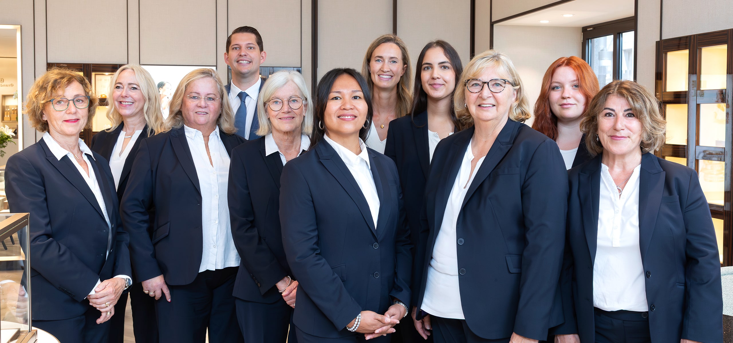 © Juwelier Wenthe Eine Gruppe von professionell gekleideten Personen in dunklen Anzügen und weißen Hemden steht in einem Innenraum zusammen und lächelt in die Kamera vor dem Ladengeschäft.