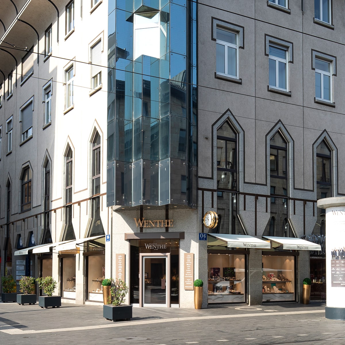 © Juwelier Wenthe Eine moderne Gebäudeecke mit Glasscheiben und geometrischen Fenstermotiven. Darunter befindet sich der Eingang zum Juweliergeschäft mit dem Namen "WENTHE", umrahmt von goldenen Akzenten und flankiert von grünen Topfpflanzen auf dem Gehweg.