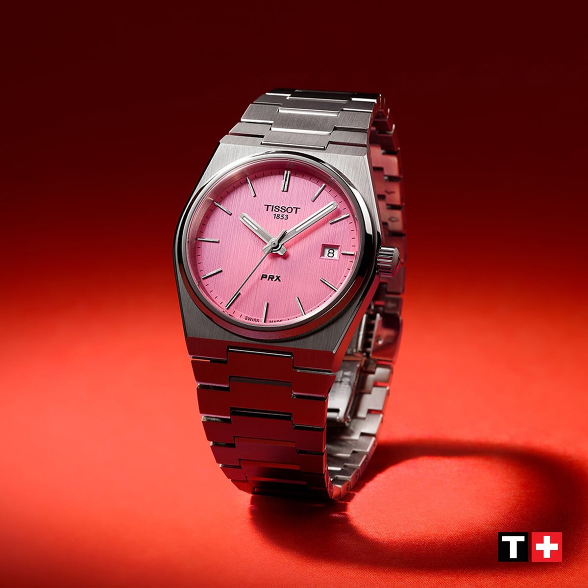 2025 © TISSOT Eine Tissot Classic Armbanduhr aus Edelstahl mit rosafarbenem Zifferblatt und Datumsanzeige wird auf rotem Hintergrund präsentiert, wobei das Tissot-Logo in der rechten unteren Ecke sichtbar ist.