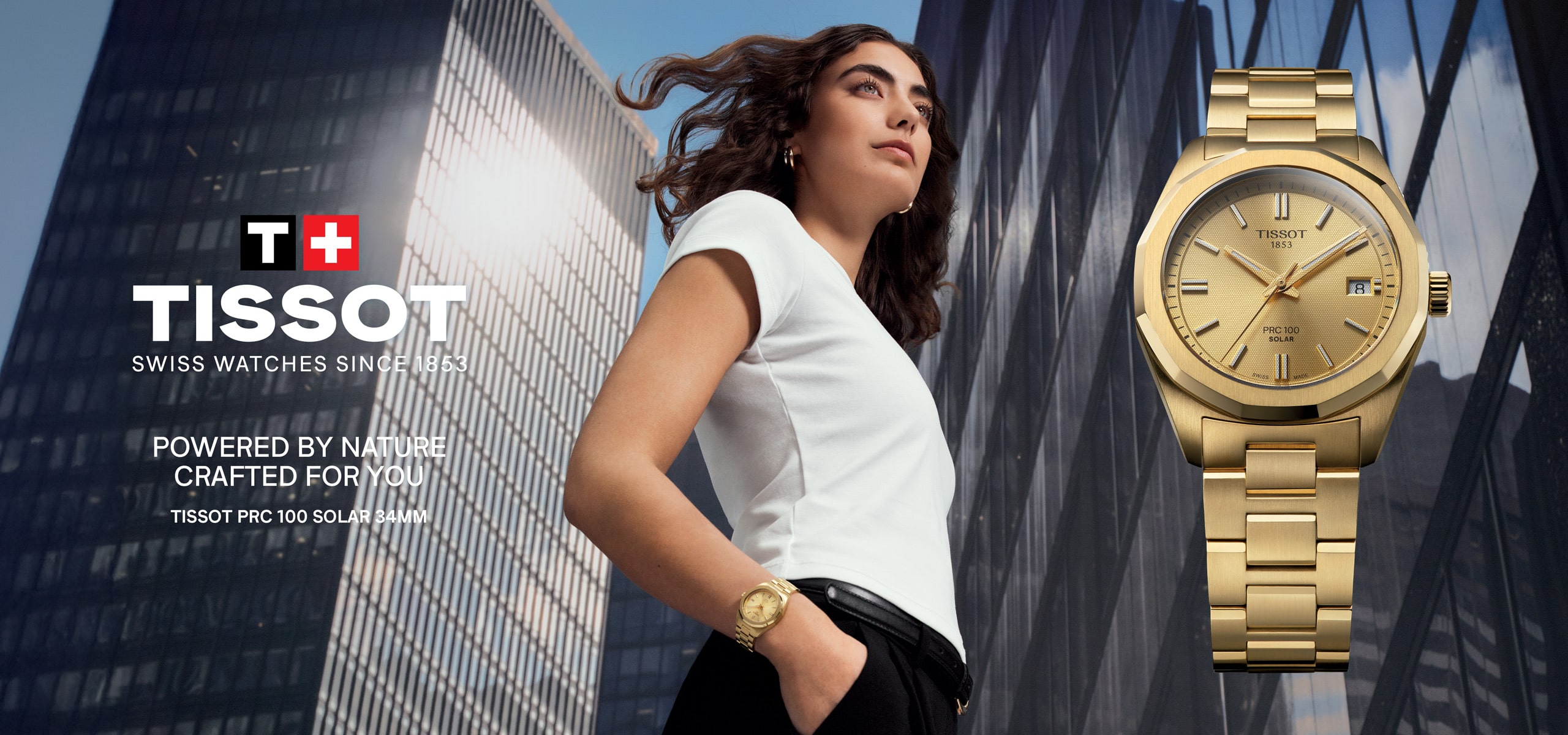2025 © TISSOT Eine Frau in einem weißen Oberteil und einer goldenen Uhr steht selbstbewusst vor modernen Glasgebäuden. Neben ihr ist eine Nahaufnahme einer goldenen Tissot PRX 100 Solaruhr zu sehen, die das Tissot-Logo und den Slogan trägt.