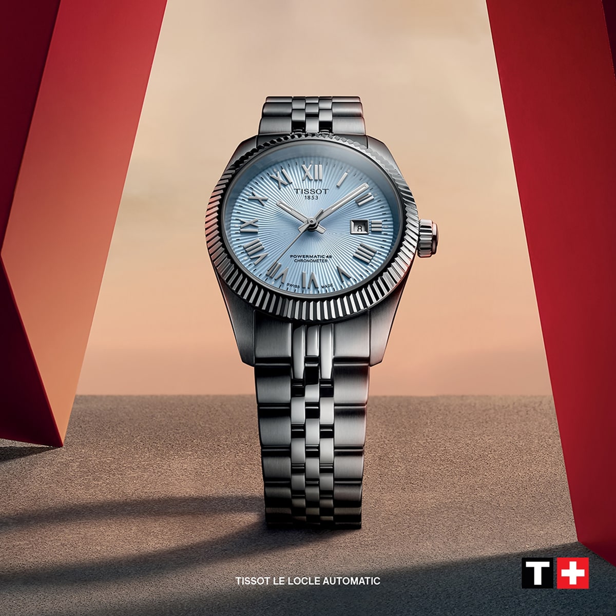 2025 © TISSOT Eine silberne Tissot Le Locle Automatikuhr mit strukturiertem hellblauem Zifferblatt steht aufrecht auf einer grauen Fläche, eingerahmt von zwei roten Strukturen vor einem beige-rosa Himmel. In der rechten unteren Ecke ist das Logo von Tissot Le Locle zu sehen.