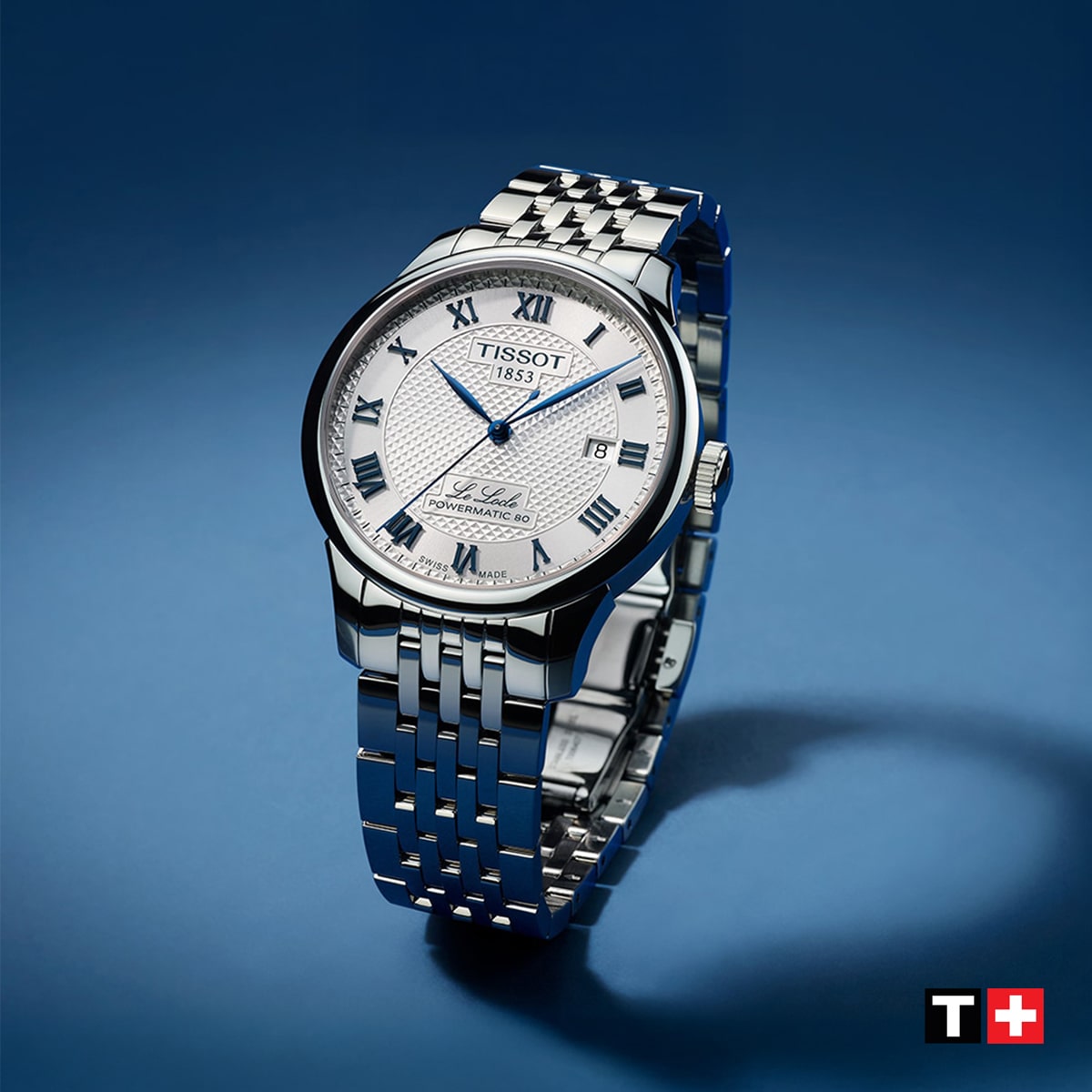 2025 © TISSOT Eine silberne Tissot Le Locle Armbanduhr mit weißem Zifferblatt, blauen römischen Ziffern und einem Metallarmband ist auf einem blauen Hintergrund mit Farbverlauf abgebildet. Das Tissot-Logo erscheint in der rechten unteren Ecke.