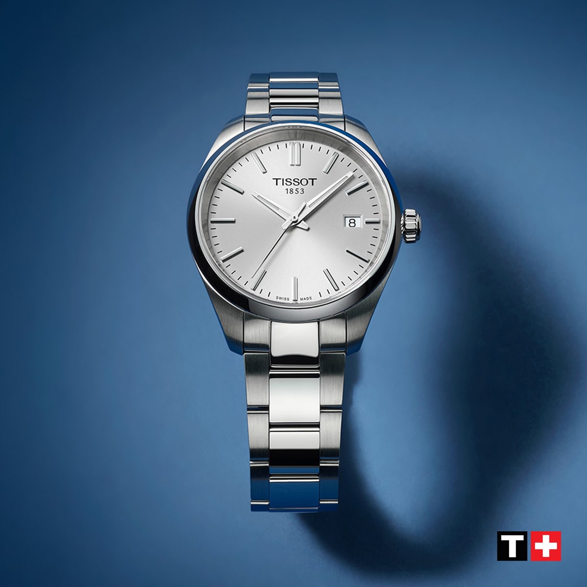 2025 © TISSOT Eine silberne Tissot PR 100 Armbanduhr mit einem Metallband ist auf einem blauen Hintergrund abgebildet. Die Uhr hat ein schlichtes, elegantes Zifferblatt mit einem Datumsfenster. Das Tissot-Logo und ein rotes Schweizerkreuz erscheinen in der unteren rechten Ecke.