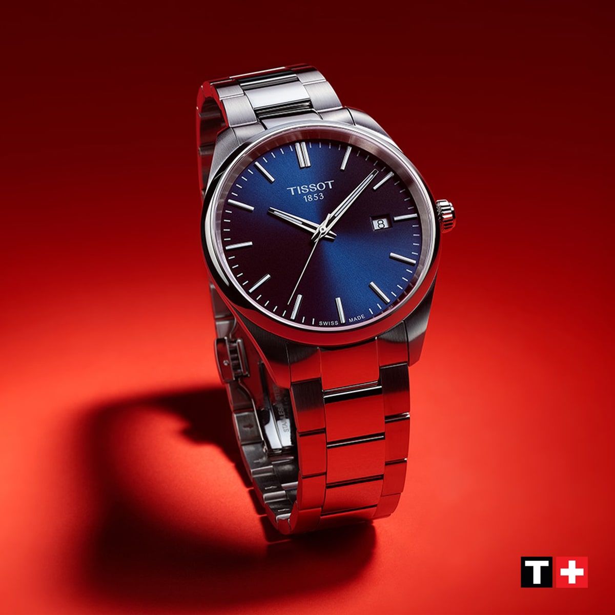 2025 © TISSOT Eine Tissot PR 100 Armbanduhr mit blauem Zifferblatt und silbernem Metallband ist aufrecht auf einem roten Hintergrund abgebildet. Das Datum wird bei 3 Uhr angezeigt und das Tissot-Logo erscheint in der rechten unteren Ecke.