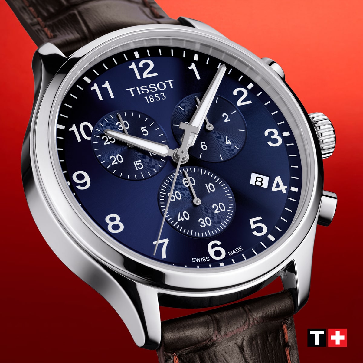 2025 © TISSOT Nahaufnahme einer Tissot XL-Chronographenuhr mit blauem Zifferblatt, silbernen Zeigern, drei Hilfszifferblättern, Datumsfenster und braunem Lederarmband vor einem roten Hintergrund mit dem Tissot-Logo in der Ecke.