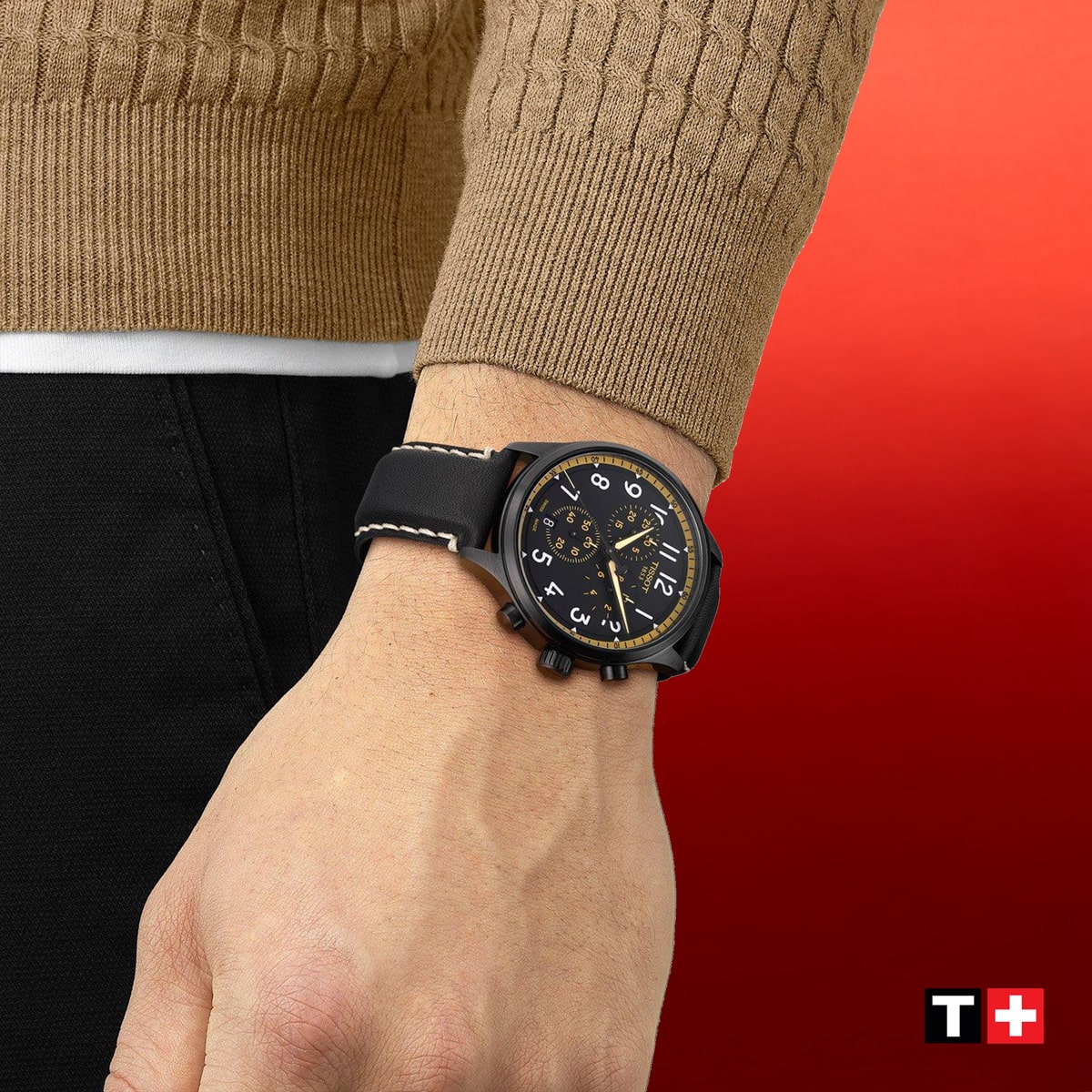 2025 © TISSOT Eine Person, die einen braunen Pullover und eine schwarze Hose trägt, zeigt eine Tissot XL-Armbanduhr mit schwarz-gelbem Zifferblatt. Der Hintergrund ist rot, und das Tissot-Logo erscheint in der unteren rechten Ecke.