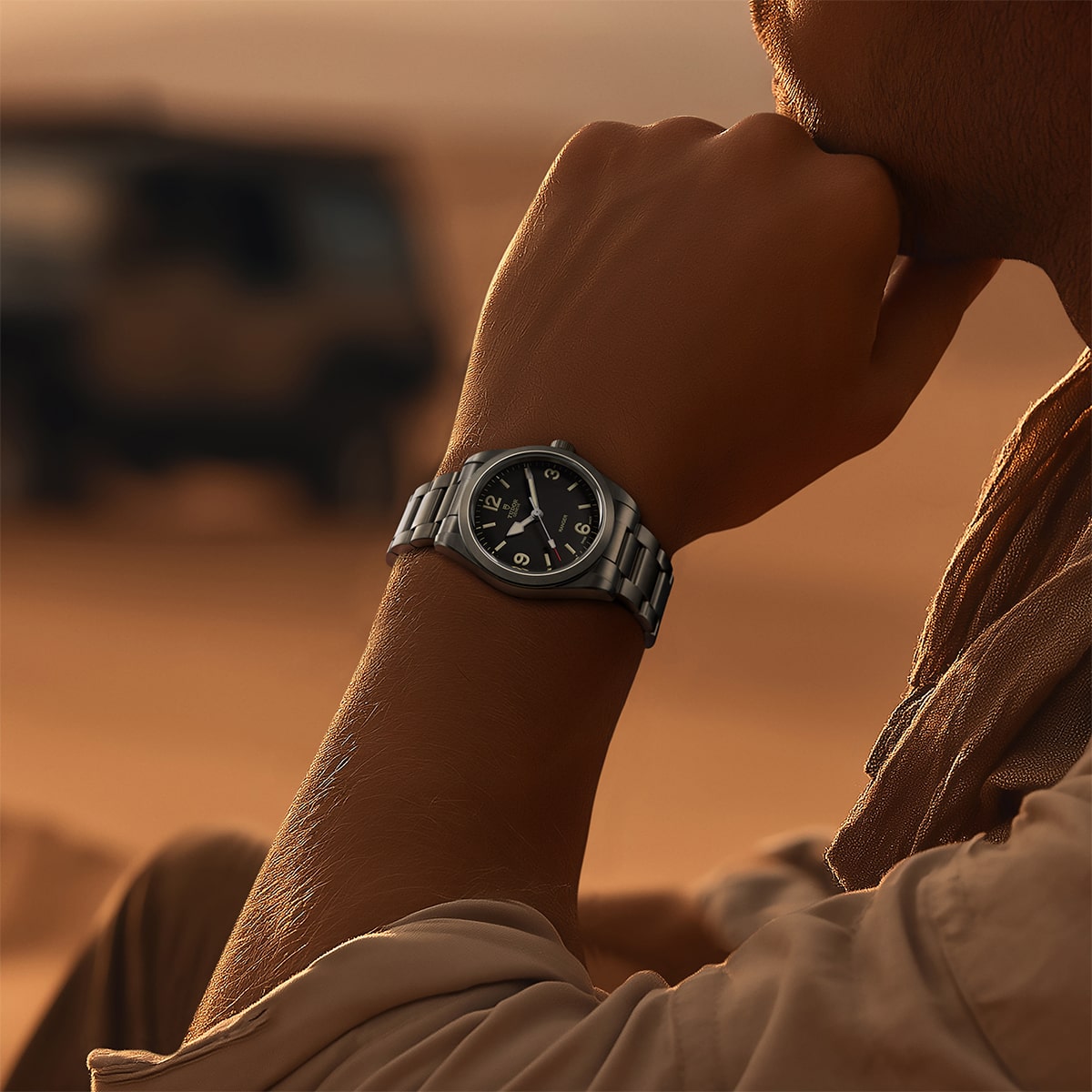 2026 © TUDOR Eine Person, die eine silberne Tudor Ranger-Armbanduhr mit schwarzem Zifferblatt trägt, sitzt im Freien in einer Wüstenlandschaft und stützt ihr Kinn auf die Hand. Im sandigen Hintergrund ist ein unscharfes Fahrzeug zu erkennen.