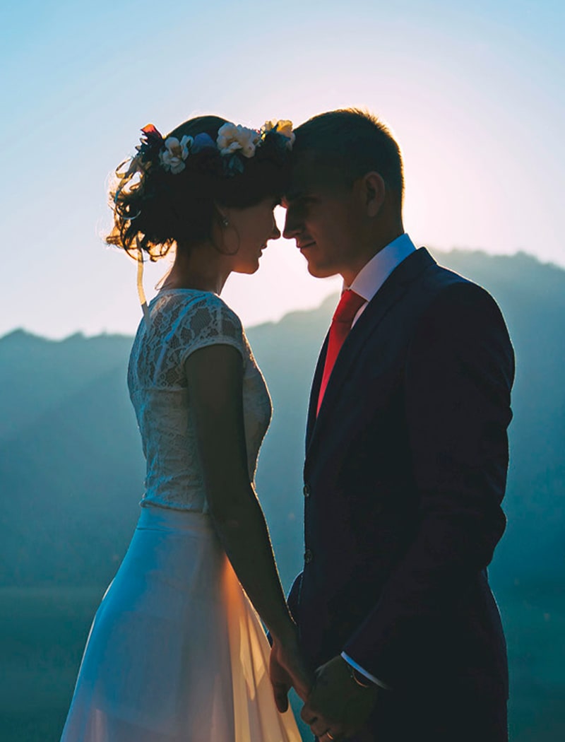 2025 © 123GOLD Eine Braut und ein Bräutigam stehen bei Sonnenuntergang im Freien dicht beieinander, berühren sich an den Stirnen und halten sich an den Händen. Die Braut trägt eine Blumenkrone und ein Spitzenkleid, während der Bräutigam einen Anzug, eine rote Krawatte und einen 123GOLD-Ehering trägt. Im Hintergrund sind Berge zu sehen.