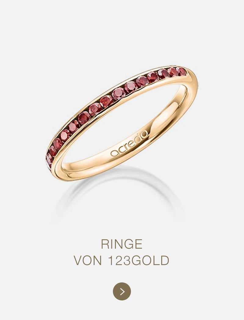 Ein schmaler Goldring, besetzt mit einer Reihe kleiner roter Edelsteine. Die Innenseite des Rings ist mit "acredo" graviert. Der Text darunter lautet: "RINGE VON 123GOLD - exklusiv gefertigt für einzigartige Momente.
