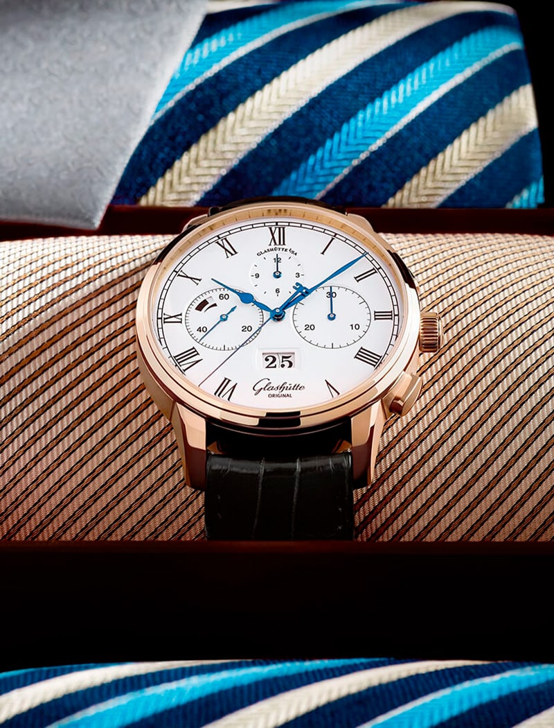 2025 © Glashütte Original Eine Glashütte Original Luxus-Armbanduhr mit weißem Zifferblatt, blauen Zeigern, römischen Ziffern und schwarzem Lederarmband wird in einer Schachtel präsentiert. Im Hintergrund sind blau und beige gestreifte Krawatten zu sehen.