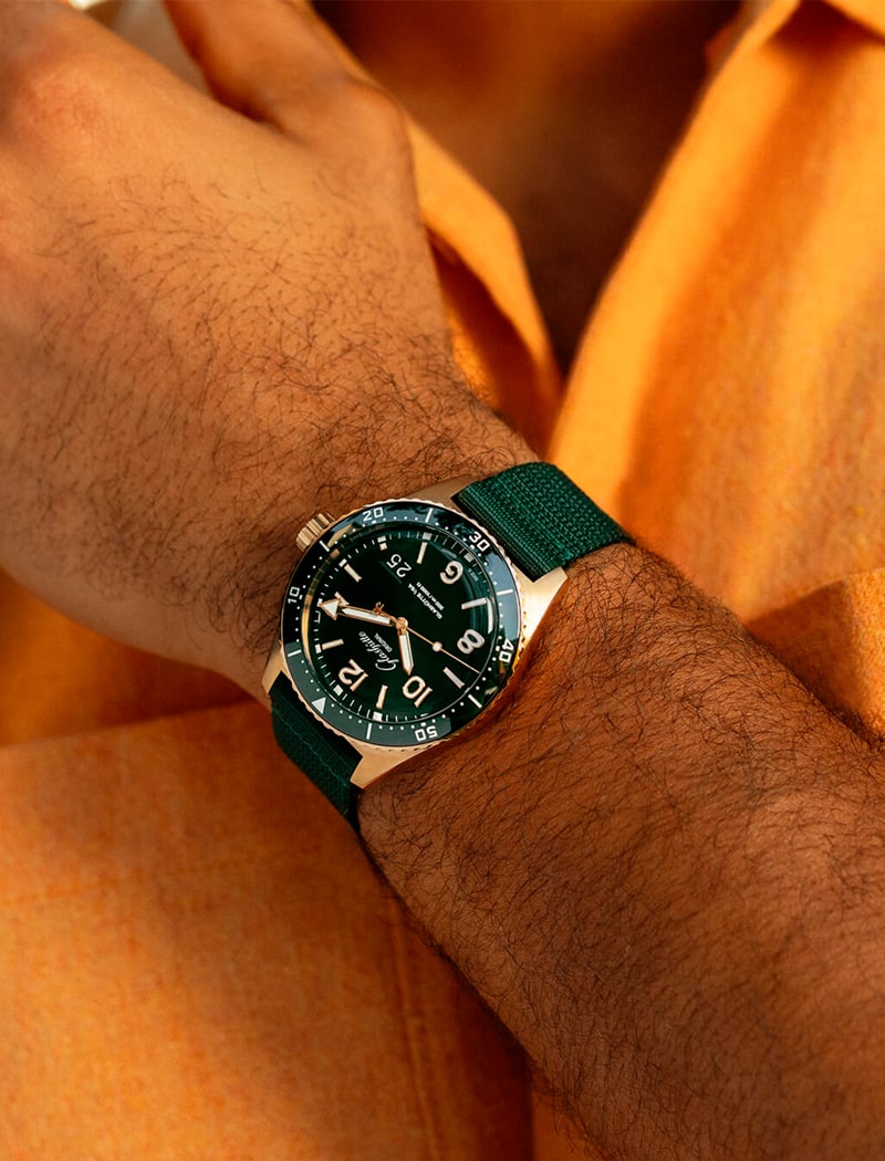 2025 © Glashütte Original Nahaufnahme des Handgelenks einer Person, die eine Glashütte Original Armbanduhr mit grünem Armband, dunkelgrünem Zifferblatt und fetten weißen Ziffern trägt, vor dem Hintergrund eines orangefarbenen Hemdes.