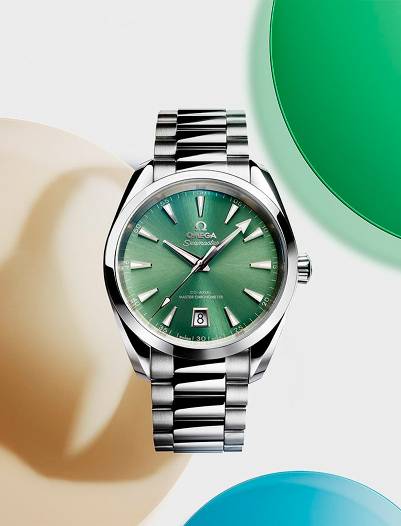 2025 © OMEGA Eine silberne Omega-Armbanduhr mit grünem Zifferblatt, Datumsanzeige und Edelstahlarmband wird vor einem weißen Hintergrund präsentiert, der durch weiche, bunte Kreisformen in Beige, Grün und Blau akzentuiert wird.