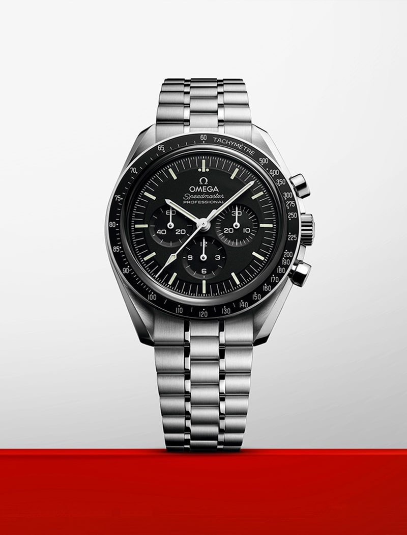2025 © OMEGA Eine Omega Speedmaster Professional Armbanduhr aus Edelstahl mit schwarzem Zifferblatt, drei Hilfszifferblättern und einem Metallarmband wird vor einem weißen Hintergrund mit einem roten Streifen am unteren Rand präsentiert.
