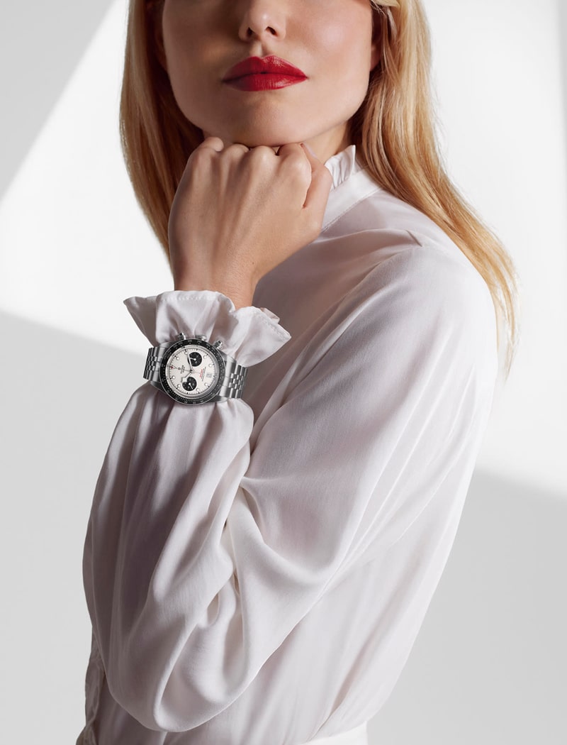 Eine Frau in weißer Bluse und mit rotem Lippenstift posiert mit der Hand in der Nähe ihres Gesichts und präsentiert eine stilvolle silberne Tudor-Armbanduhr mit rundem Zifferblatt und drei Ziffernblättern an ihrem Handgelenk.