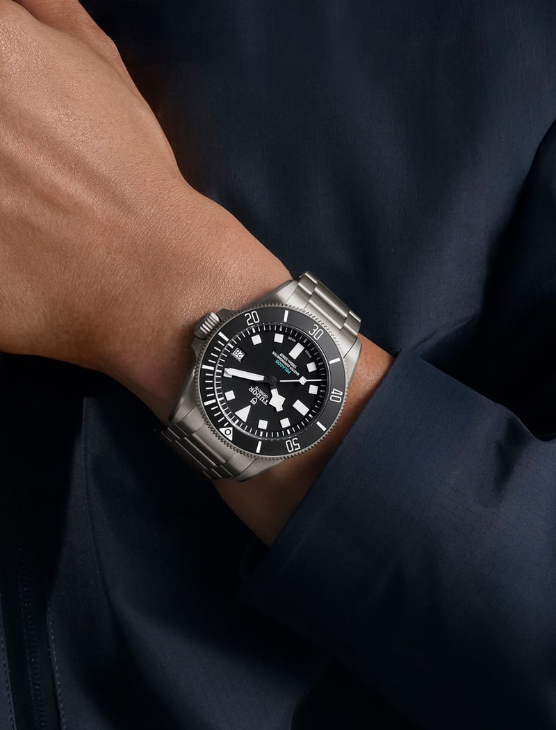 Eine Person, die eine silberne Tudor-Taucheruhr aus Edelstahl mit schwarzem Zifferblatt, auffälligen weißen Indexen und einer drehbaren Lünette trägt, wird an ihrem Handgelenk vor einem dunklen Ärmel gezeigt.