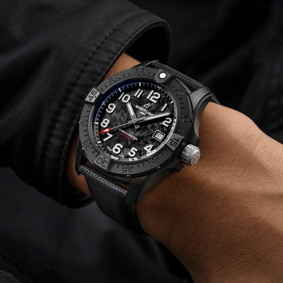 Nahaufnahme eines Handgelenks, das eine schwarze Breitling Avenger Uhr mit strukturiertem Zifferblatt, weißen Ziffern und schwarzem Armband trägt, die sich von einem dunklen Jackenärmel abhebt. Die Uhrzeiger und die Lünette sind deutlich zu erkennen.