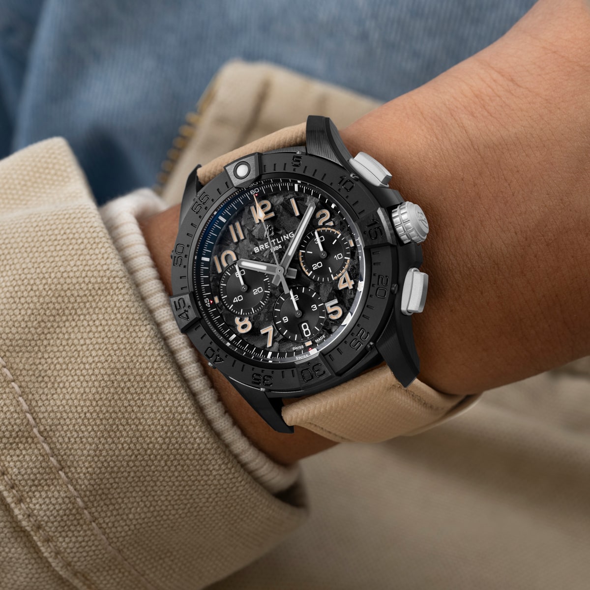 Eine Person, die eine beige Jacke und blaue Jeans trägt, trägt eine schwarze Breitling Avenger Chronographenuhr mit einem beigen Armband. Das Ziffernblatt ist mit mehreren Zifferblättern und weißen Ziffern versehen.
