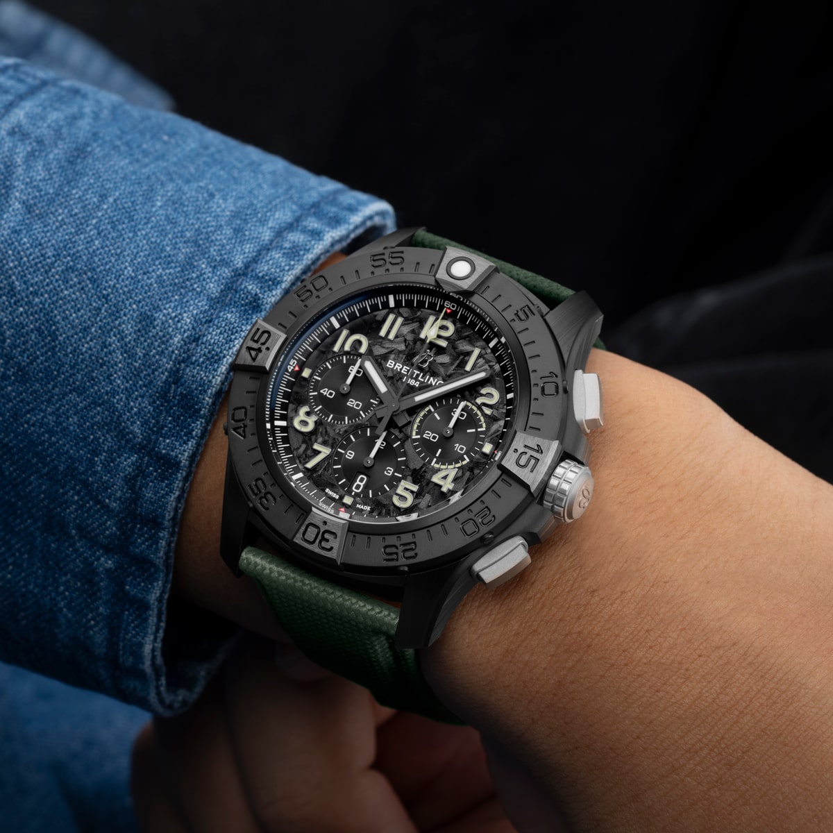 Eine Nahaufnahme einer Person, die eine schwarze Breitling Avenger Chronographenuhr mit grünem Armband trägt. Die Uhr hat ein großes, detailliertes Zifferblatt mit mehreren Ziffernblättern und einer drehbaren Lünette unter einer blauen Jeanshülle.