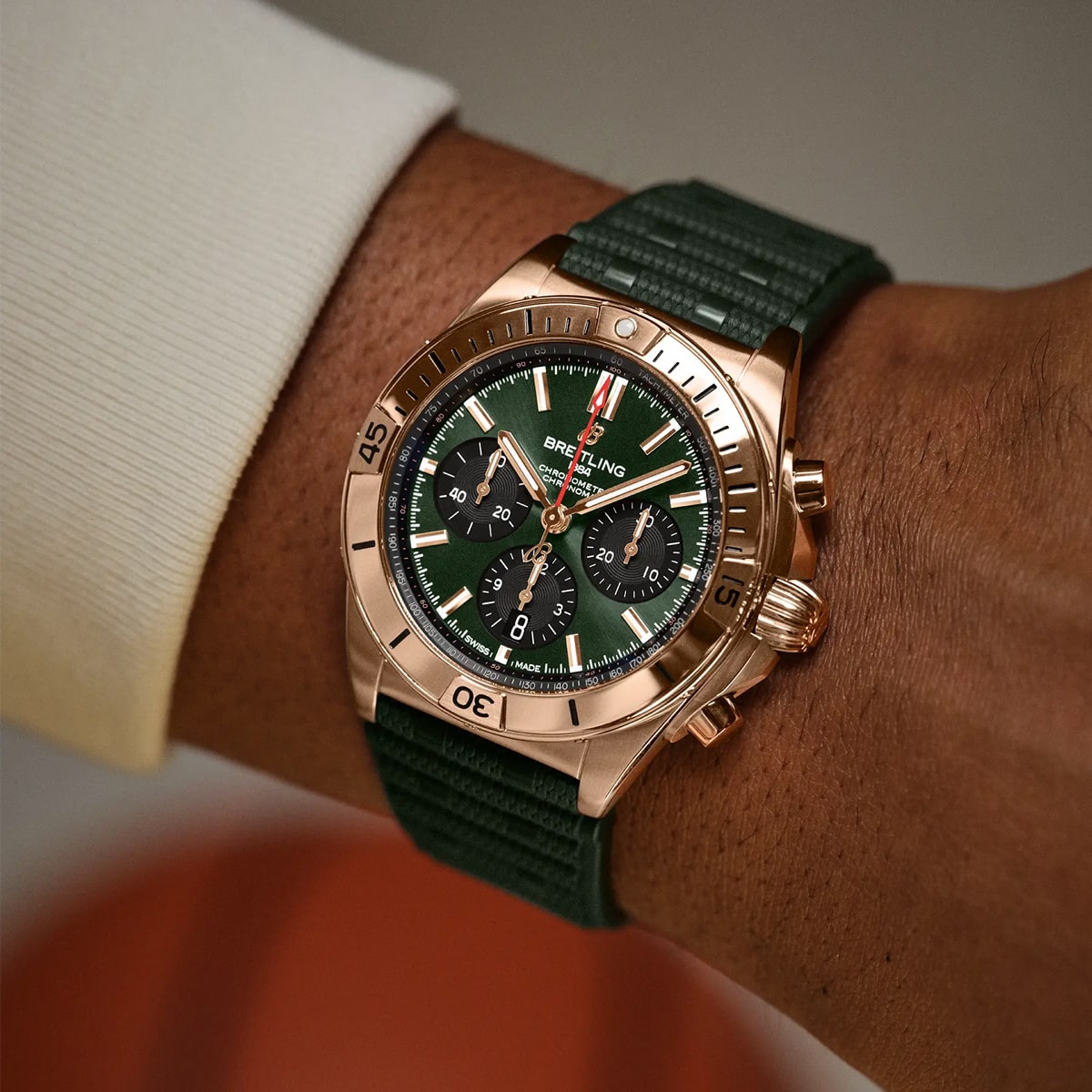 Eine Nahaufnahme des Handgelenks einer Person, die eine goldene Breitling Chronomat Chronographenuhr mit grünem Zifferblatt und passendem grünem Strukturarmband trägt. Die Person trägt ein helles, langärmeliges Hemd.