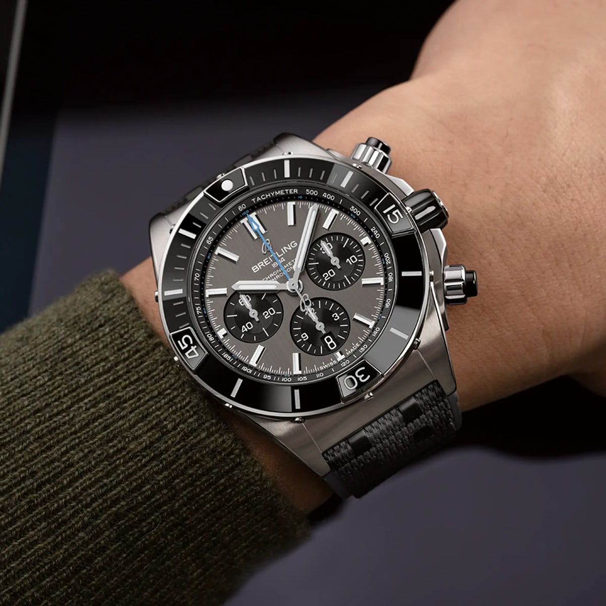 Nahaufnahme eines Handgelenks mit einer Breitling Chronomat Chronographenuhr mit schwarzer Lünette, schwarzem und grauem Zifferblatt, drei Hilfszifferblättern und schwarzem Strukturarmband. Der Träger ist mit einem grünen Pullover bekleidet.