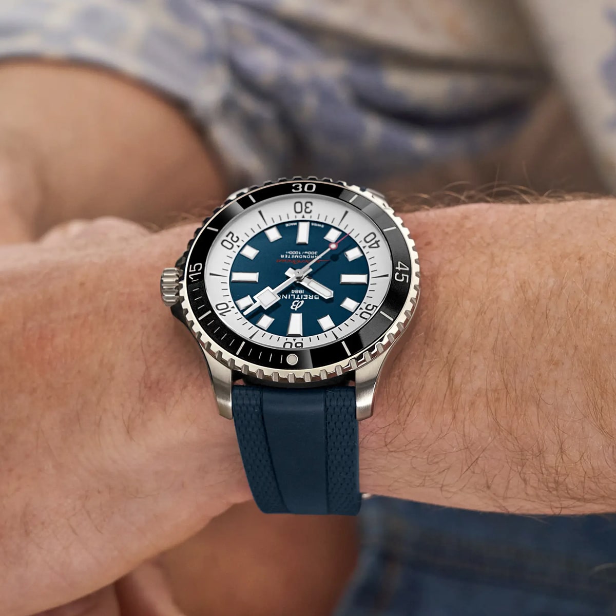 2025 © BREITLING Eine Nahaufnahme des Handgelenks einer Person, die eine Breitling Superocean-Taucheruhr mit schwarzer Lünette, silbernem Gehäuse und blauem Kautschukarmband trägt. Das Zifferblatt hat weiße Markierungen und ein Datumsfenster, und die Person trägt ein gemustertes Hemd.