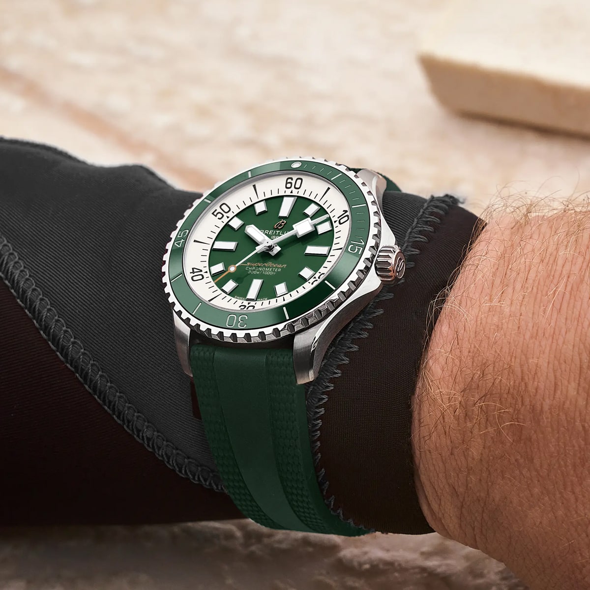 2025 © BREITLING Eine Person trägt eine grün-silberne Breitling Superocean mit einem grünen Kautschukarmband. Die Uhr verfügt über eine drehbare Lünette, auffällige weiße Stundenmarkierungen und ist mit einem schwarzen, strukturierten langen Ärmel gepaart.