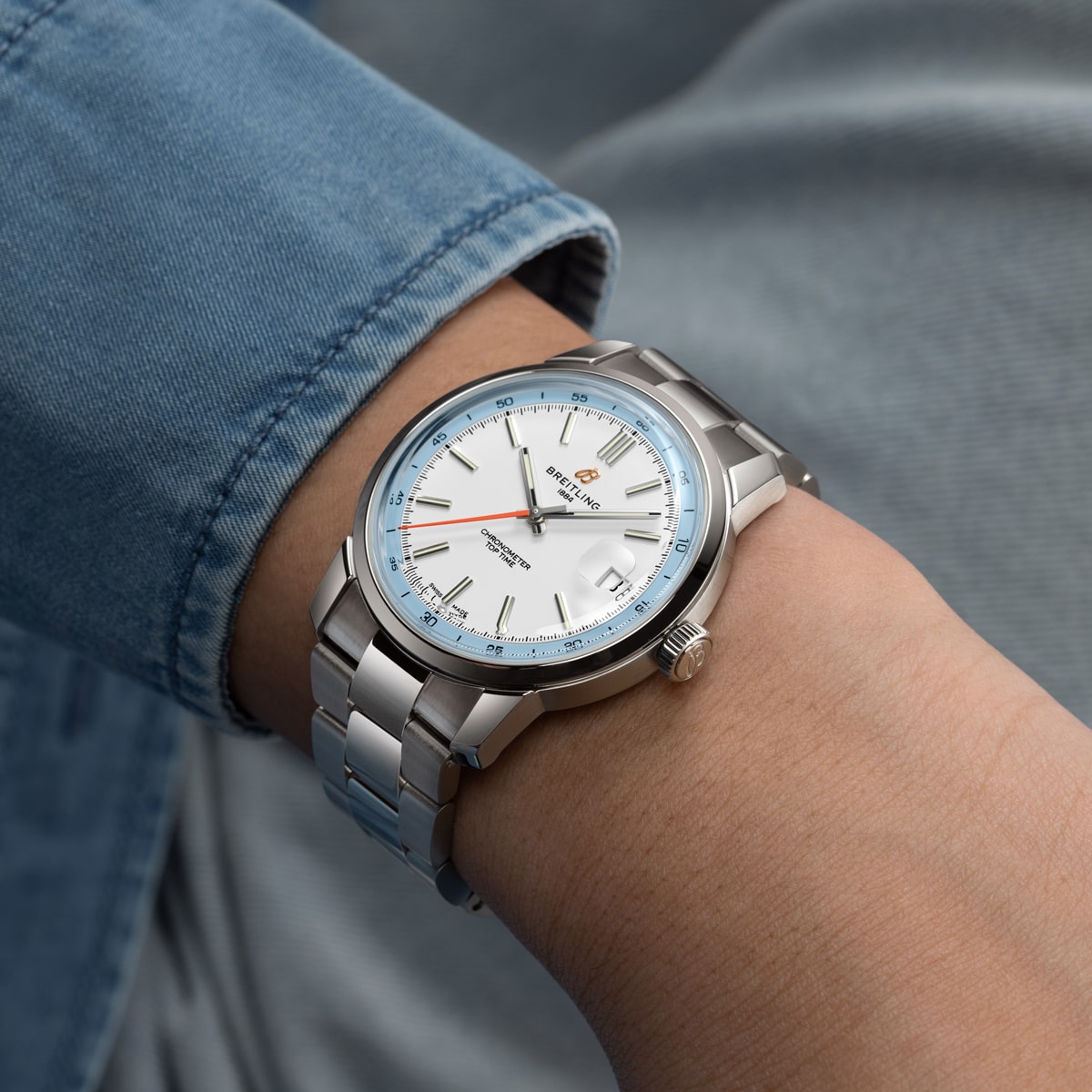2025 © BREITLING Eine Person trägt eine Breitling Top Time Edelstahl-Armbanduhr mit weißem Zifferblatt, blauen Akzenten und einem silbernen Metallband. Die Person ist mit hellblauer Jeanskleidung bekleidet.