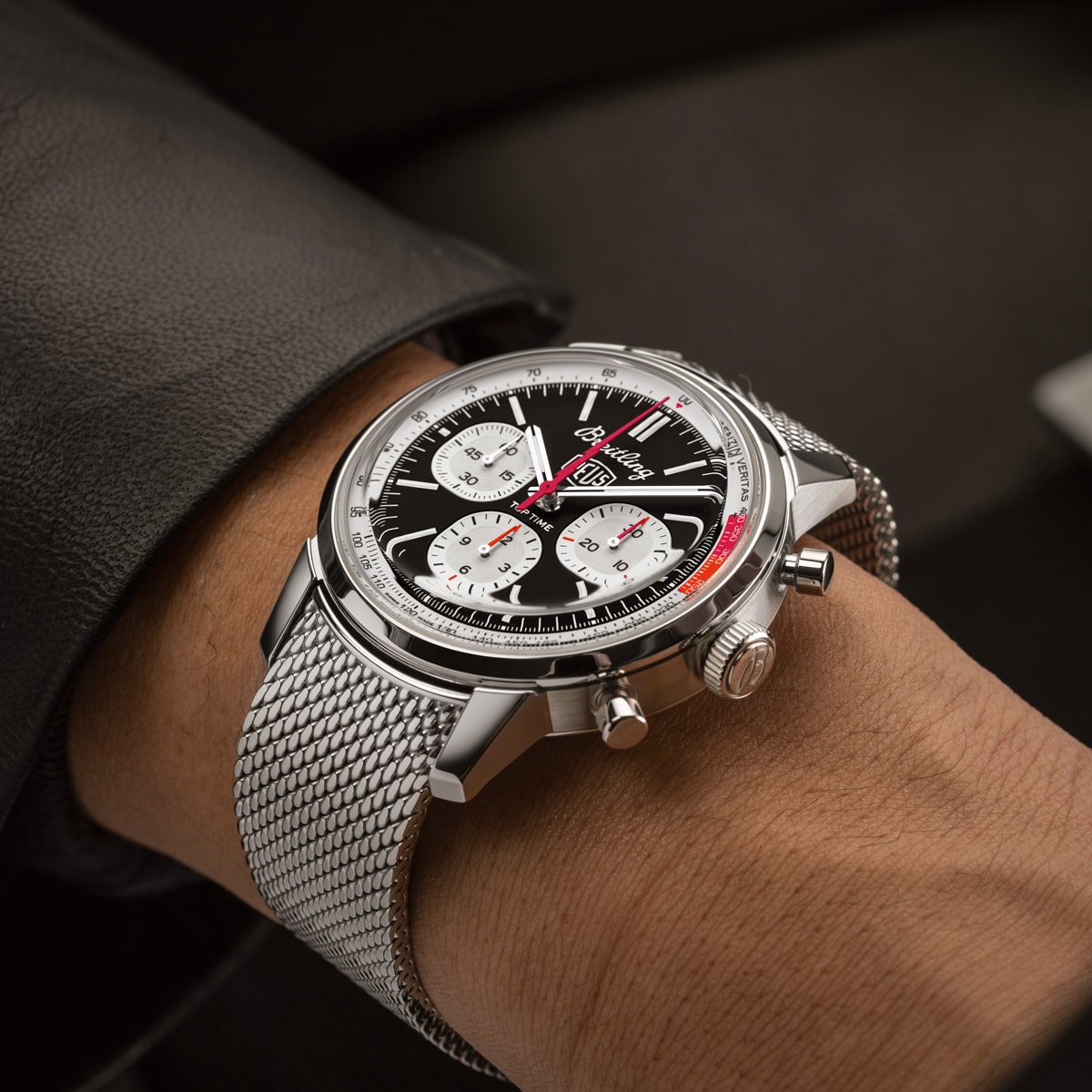 2025 © BREITLING Nahaufnahme des Handgelenks einer Person, die eine Breitling Top Time Chronographenuhr mit schwarzem Zifferblatt, weißen Hilfszifferblättern, roten Akzenten und einem Metallnetzarmband trägt. Die Person ist dunkel gekleidet.