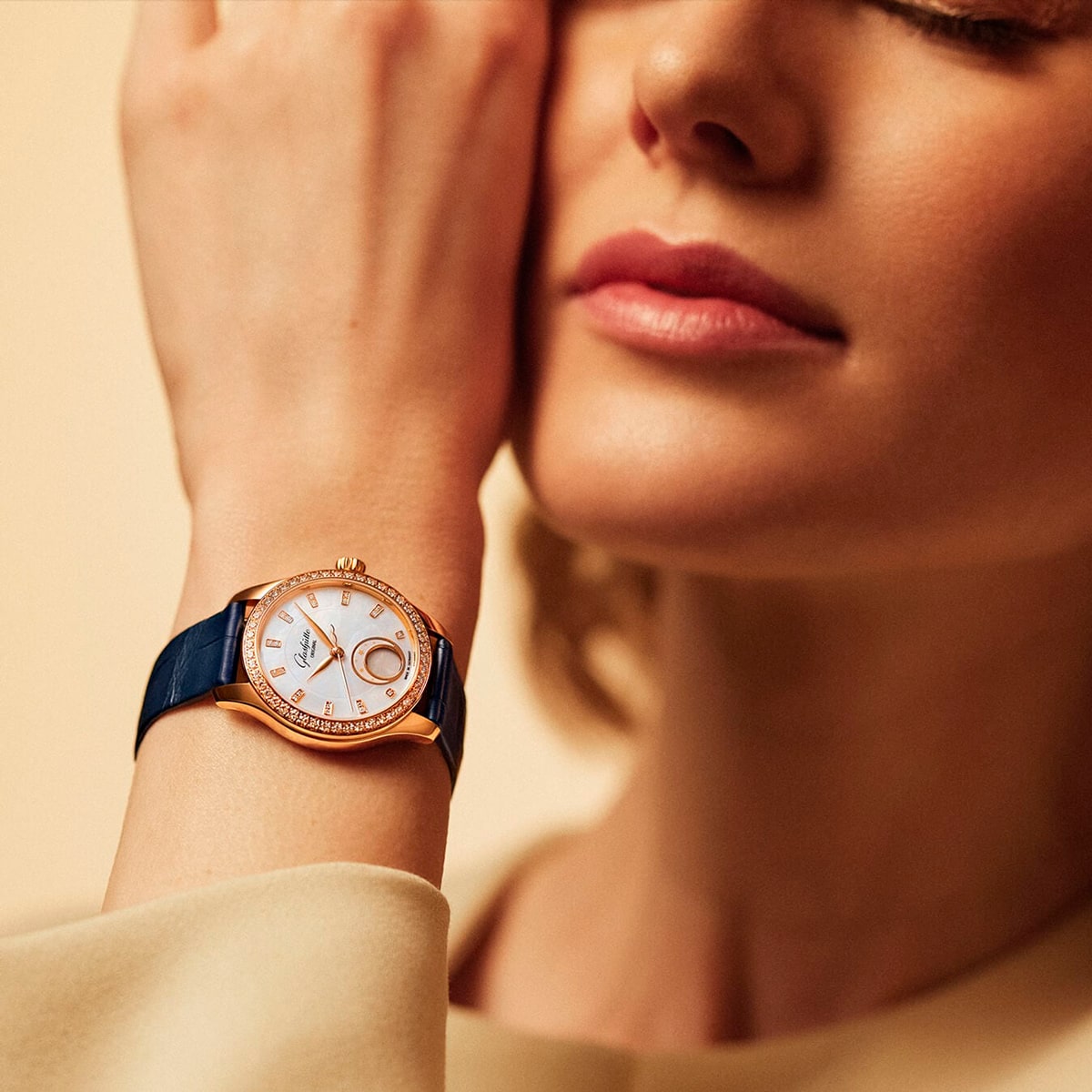 2025 © Glashütte Original Eine Frau mit geschlossenen Augen berührt ihr Gesicht und präsentiert eine goldene Glashütte Original Damenuhr mit blauem Armband und diamantbesetzter Lünette an ihrem Handgelenk. Der sanft verschwommene Hintergrund unterstreicht die Eleganz der Uhr.