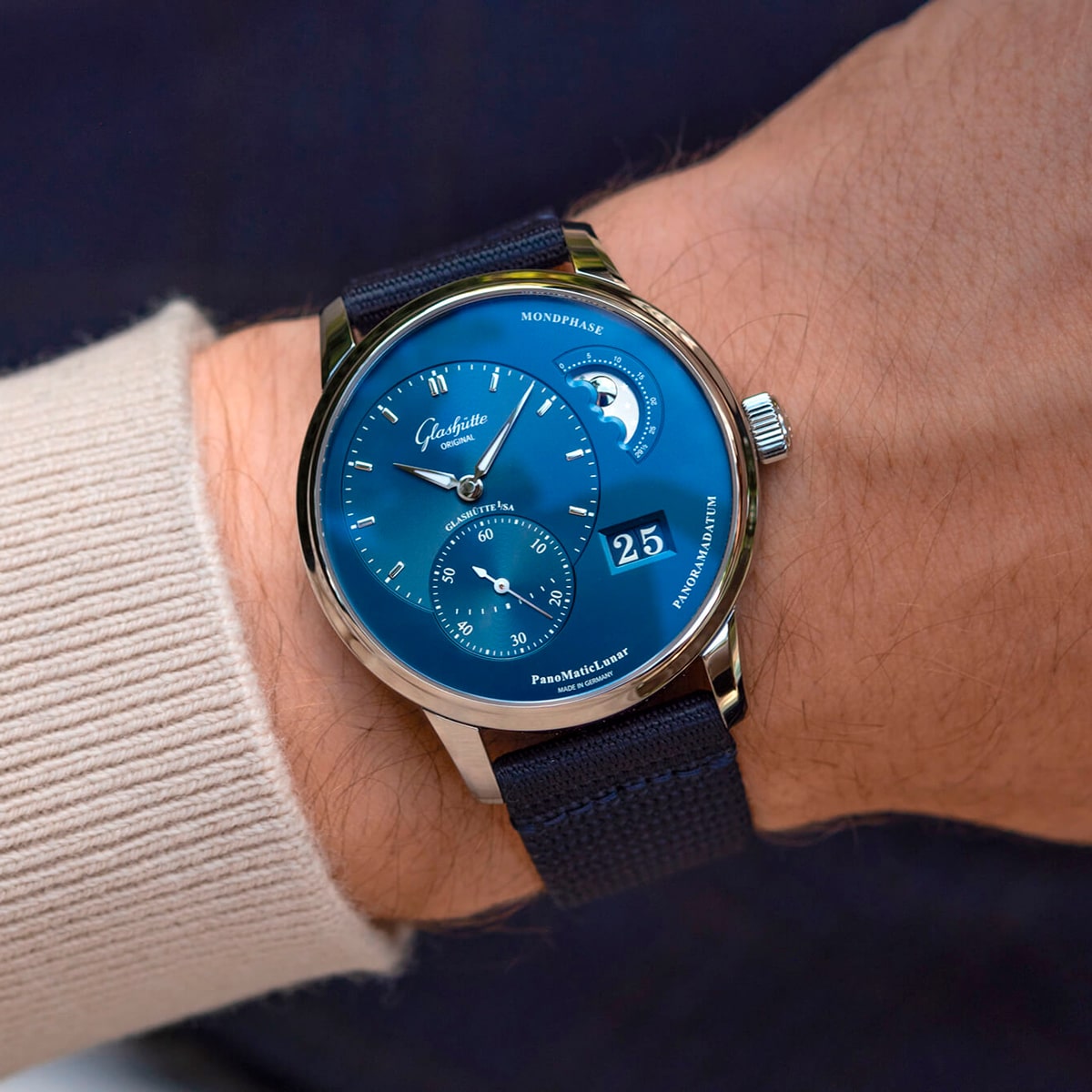 2025 © Glashütte Original Eine Nahaufnahme des Handgelenks einer Person, die eine blaue Glashütte Original Pano Kollektion Armbanduhr mit mehreren Zifferblättern, einer Mondphasenanzeige und einem marineblauen Stoffarmband trägt. Die Person trägt ein beigefarbenes langärmeliges Hemd.