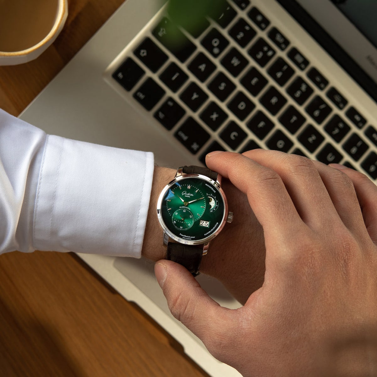2025 © Glashütte Original Eine Person mit weißem Hemd prüft eine grüngesichtige Armbanduhr der Pano-Kollektion, während sie mit einem Laptop und einer Tasse Kaffee an einem Schreibtisch sitzt.