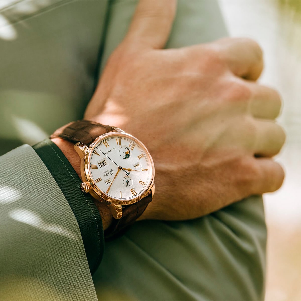 Nahaufnahme des Handgelenks einer Person, die eine goldene Senator Kollektion-Armbanduhr mit einem braunen Lederarmband trägt. Die Person ist mit einer hellgrünen Anzugsjacke bekleidet, und das Sonnenlicht schafft eine sanfte, natürliche Atmosphäre.