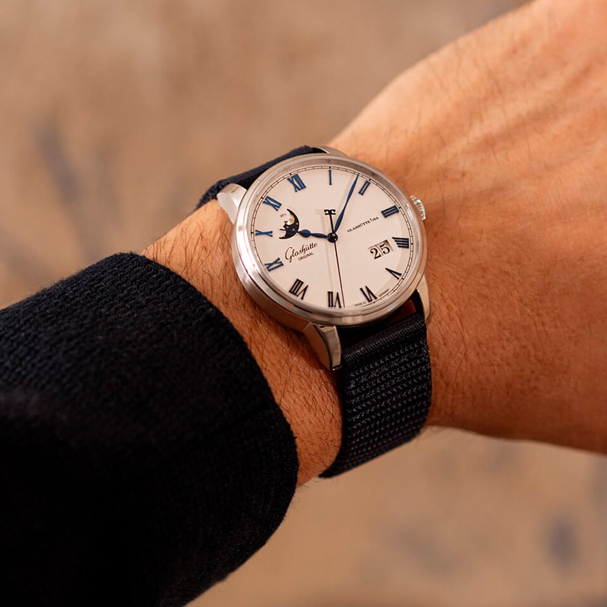 Eine Person trägt eine silberne Glashütte Original Senator Kollektion Armbanduhr mit weißem Zifferblatt, blauen Zeigern, römischen Ziffern, Mondphasen- und Datumsanzeige an einem dunklen gewebten Armband.