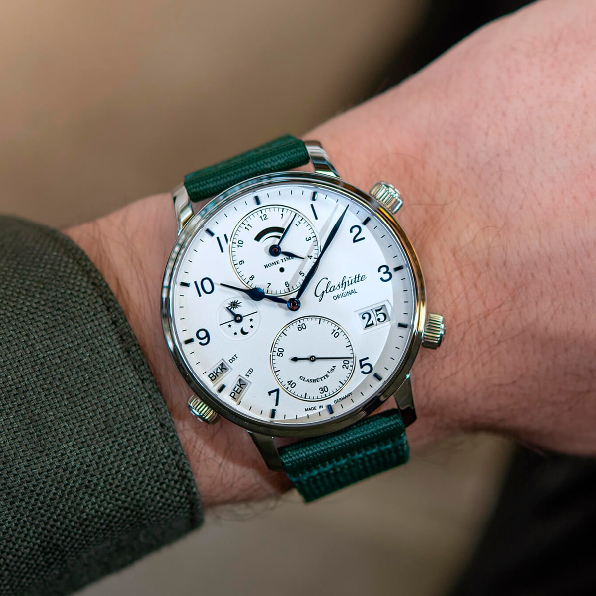 Nahaufnahme eines Handgelenks mit einer Glashütte Original Senator Kollektion Uhr mit weißem Zifferblatt, grünem Armband und mehreren Hilfszifferblättern, die verschiedene Komplikationen anzeigen, darunter eine Mondphase und das Datum. Die Person trägt einen grünen Ärmel.