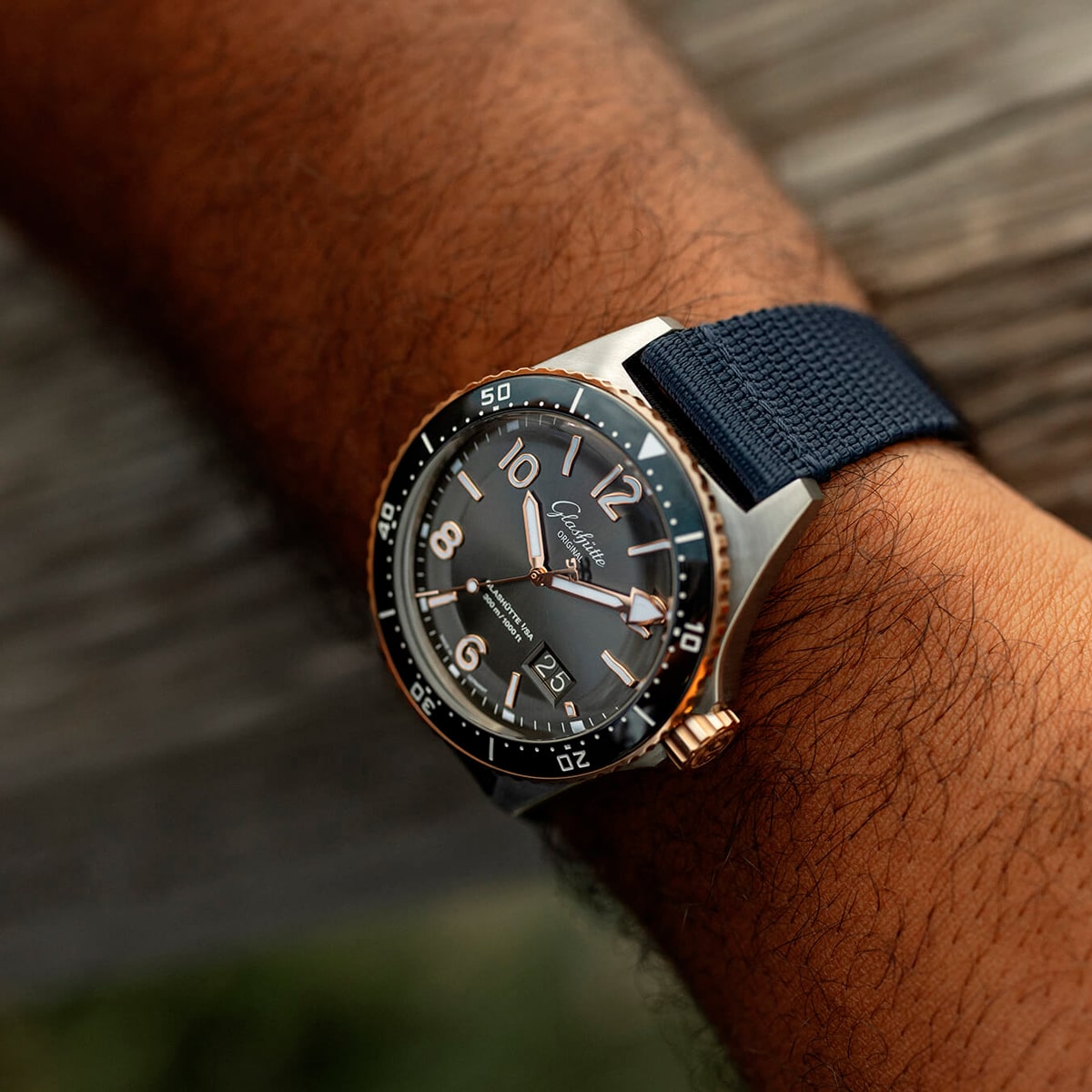 Nahaufnahme eines Handgelenks, das eine Luxusuhr der Spezialist Kollektion trägt, mit schwarzem Zifferblatt, großen silbernen und goldenen Indexen, einer drehbaren Lünette und einem blauen Stoffarmband. Der Hintergrund ist unscharfes Holz.