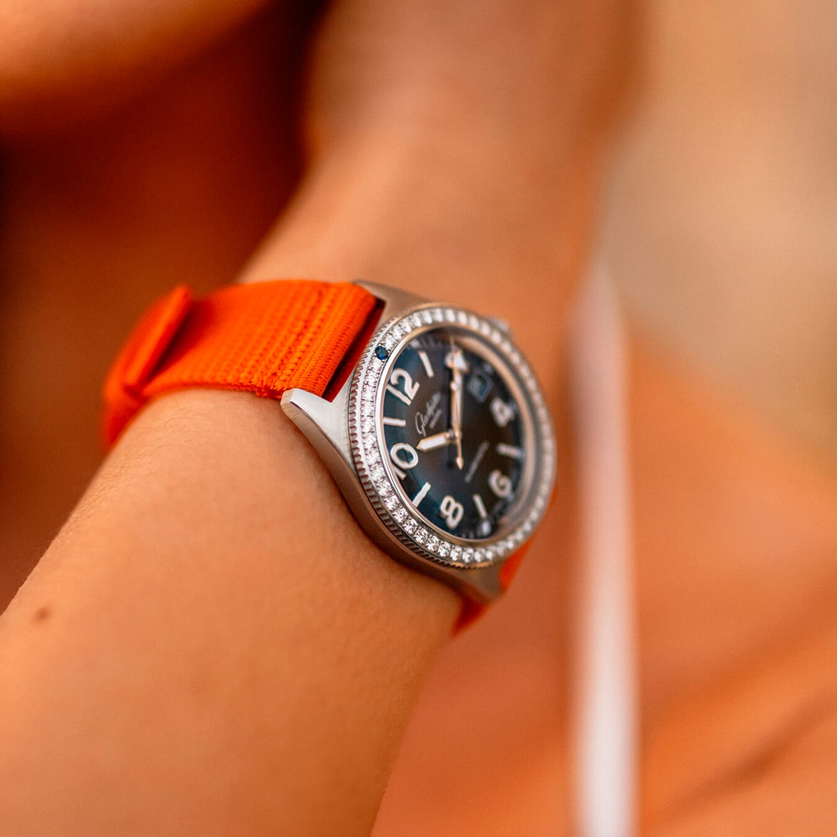 Nahaufnahme des Handgelenks einer Person, die eine Uhr der Spezialist Kollektion mit schwarzem Zifferblatt, großen weißen Ziffern, einer diamantbesetzten Lünette und einem leuchtend orangefarbenen Stoffarmband trägt. Der Hintergrund ist sanft in warmen Tönen verschwommen.