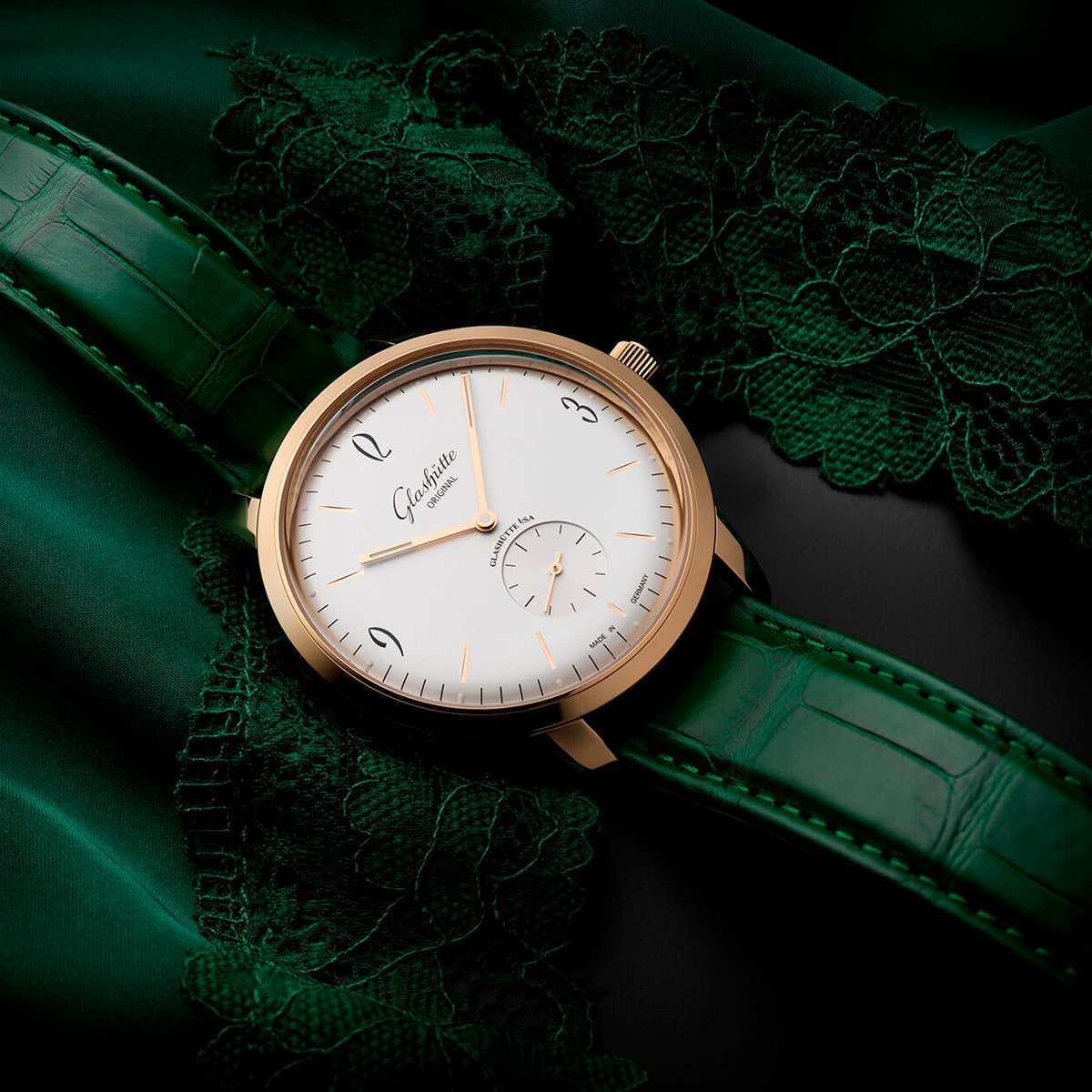 Eine goldene Armbanduhr aus der Vintage-Kollektion mit weißem Zifferblatt, schlanken schwarzen Indexen und einem grünen Lederarmband ruht auf smaragdgrünem Spitzenstoff. Die Uhr trägt den Markennamen "Glashütte Original".