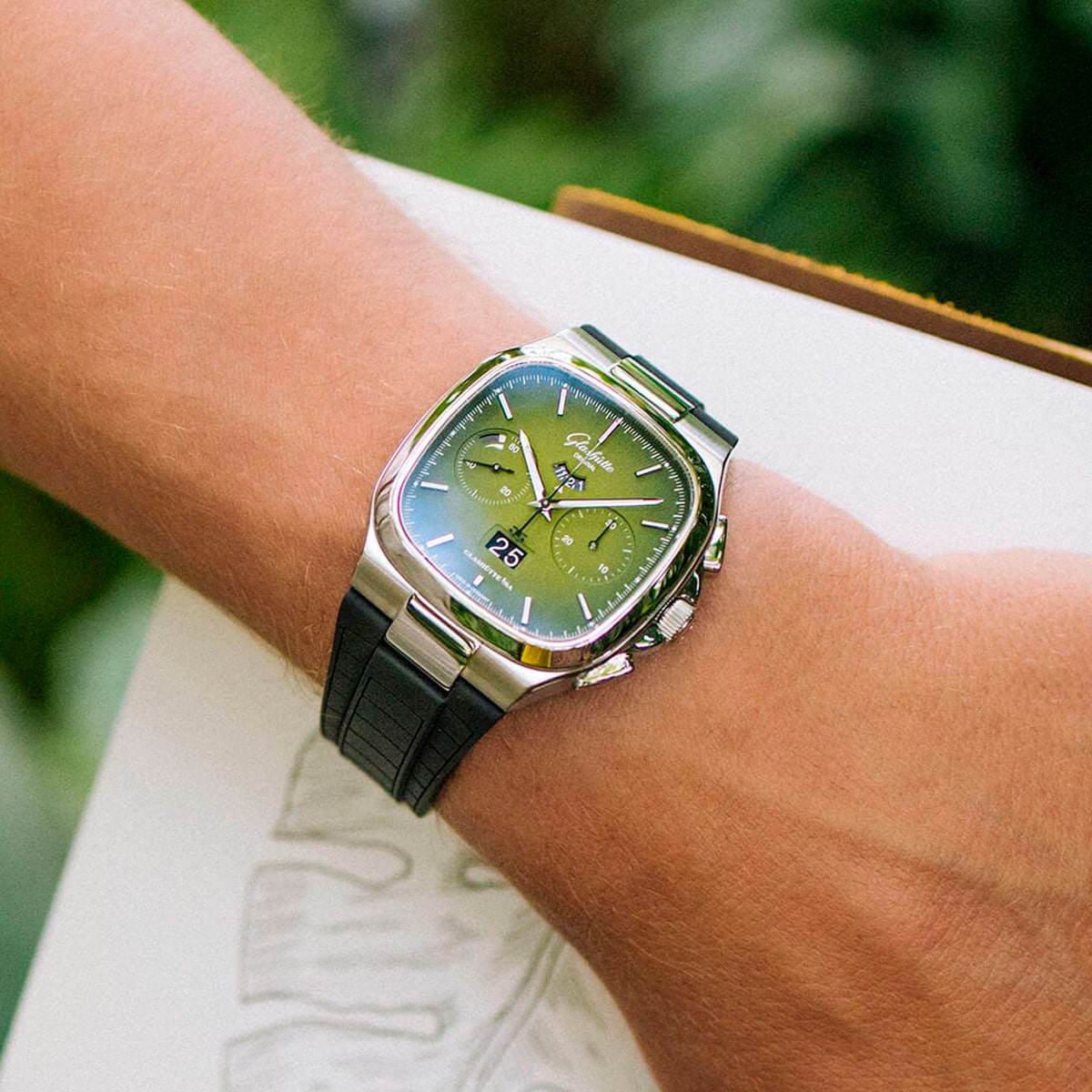 Eine Person trägt eine silberne Vintage-Kollektion-Uhr mit grünem Zifferblatt und schwarzem Krokodillederarmband am Handgelenk und hält ein Buch mit schwachen Skizzen; im Hintergrund ist grünes Laub zu sehen.