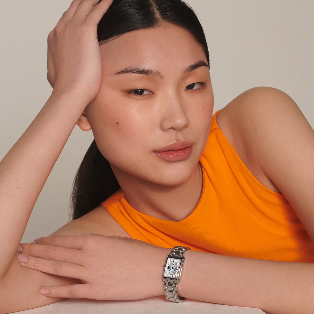 Eine Frau mit glattem schwarzem Haar, die ein orangefarbenes ärmelloses Oberteil trägt, stützt ihren Kopf auf ihre Hand und lehnt sich an einen Tisch, wobei sie eine silberne DolceVita-Armbanduhr mit rechteckigem Zifferblatt an ihrem linken Handgelenk zur Schau stellt.