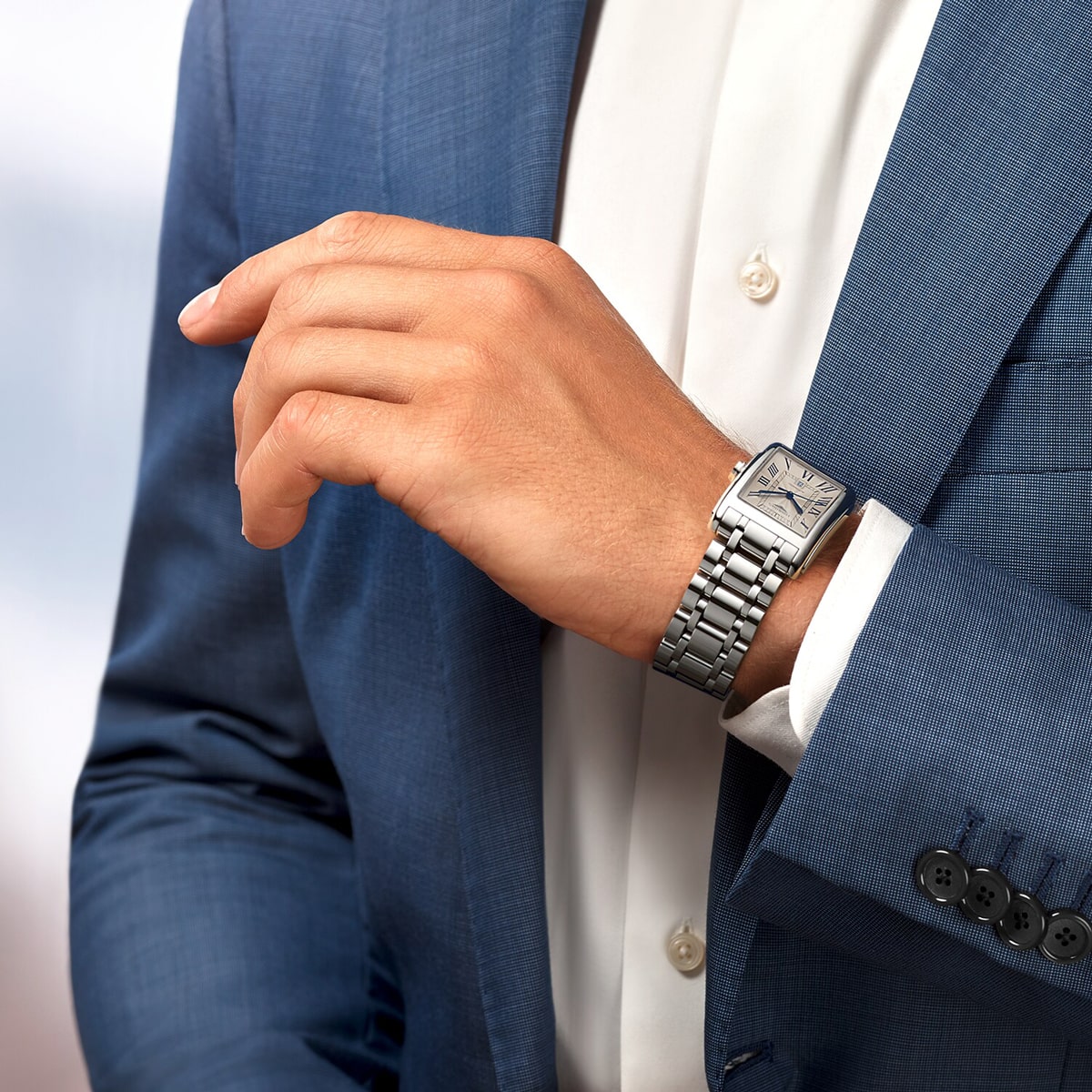 Eine Person, die einen blauen Anzug und ein weißes Hemd trägt, rückt ihren Ärmel zurecht und präsentiert eine silberne DolceVita-Armbanduhr mit rechteckigem Zifferblatt an ihrem linken Handgelenk.