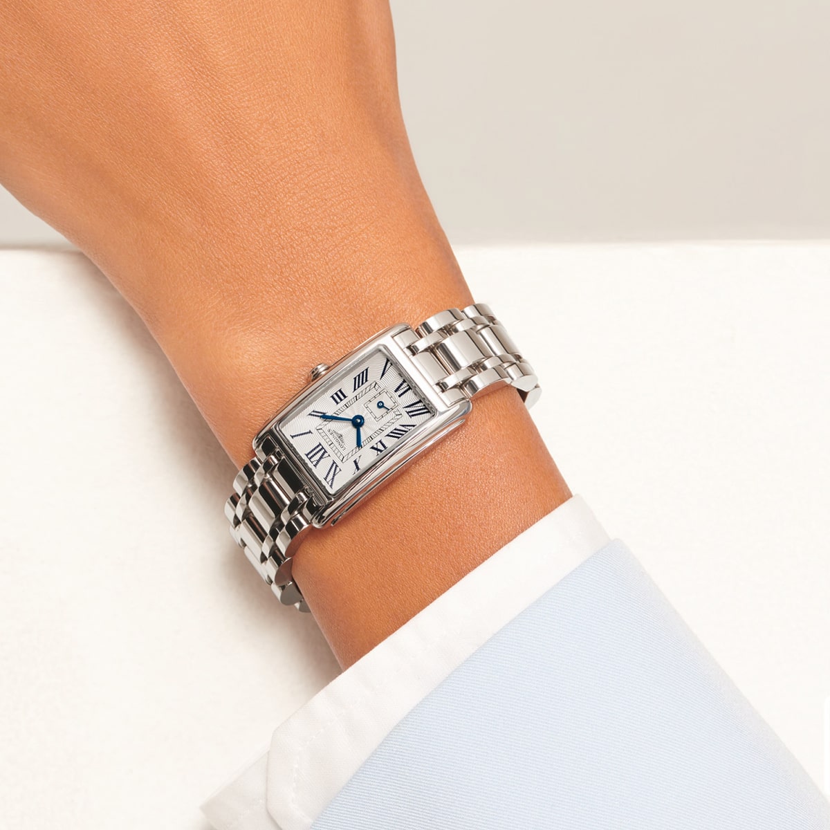 Nahaufnahme des Handgelenks einer Person, die eine rechteckige DolceVita-Silberarmbanduhr mit weißem Zifferblatt, römischen Ziffern und einem Metallgliederband trägt. Die Person ist mit einem hellblauen Hemd bekleidet.
