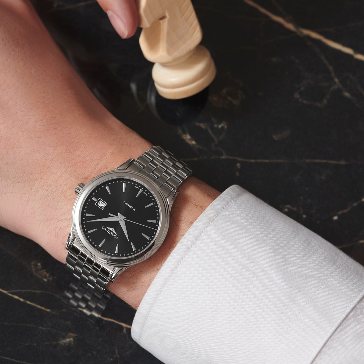 Eine Person, die eine silberne Armbanduhr mit schwarzem Zifferblatt und Metallband trägt, stützt ihre Hand auf eine schwarze Marmorfläche und hält eine hölzerne Schachfigur.