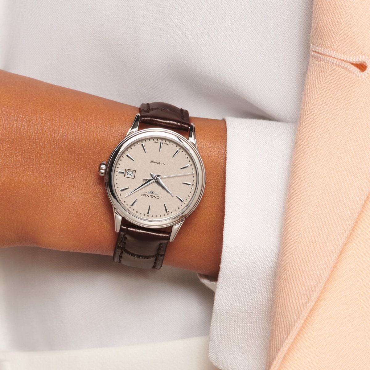 Eine Person trägt eine Armbanduhr mit braunem Lederarmband, silbernem Gehäuse und beigem Zifferblatt, die 8:22 Uhr anzeigt. Sie ist mit einem weißen Hemd und einer hellbeigen Jacke bekleidet.