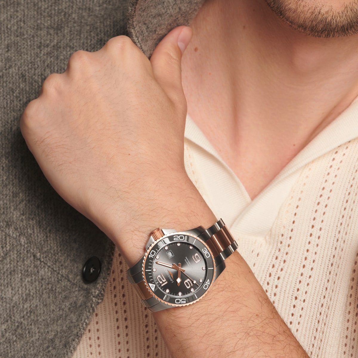 Eine Person, die eine graue und roségoldene HydroConquest-Armbanduhr mit schwarzem Zifferblatt und silbernem Band trägt, stützt ihre Hand auf ihre Brust. Die Person ist mit einem cremefarbenen Strickhemd und einer grauen, strukturierten Jacke bekleidet.