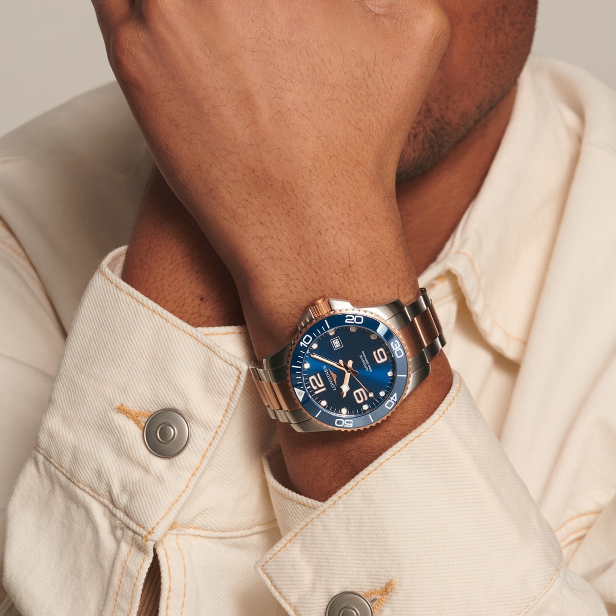 Eine Person, die eine cremefarbene Jacke trägt, hält ihren Arm hoch und zeigt eine HydroConquest-Armbanduhr in Silber und Roségold mit blauem Zifferblatt und weißen Markierungen.