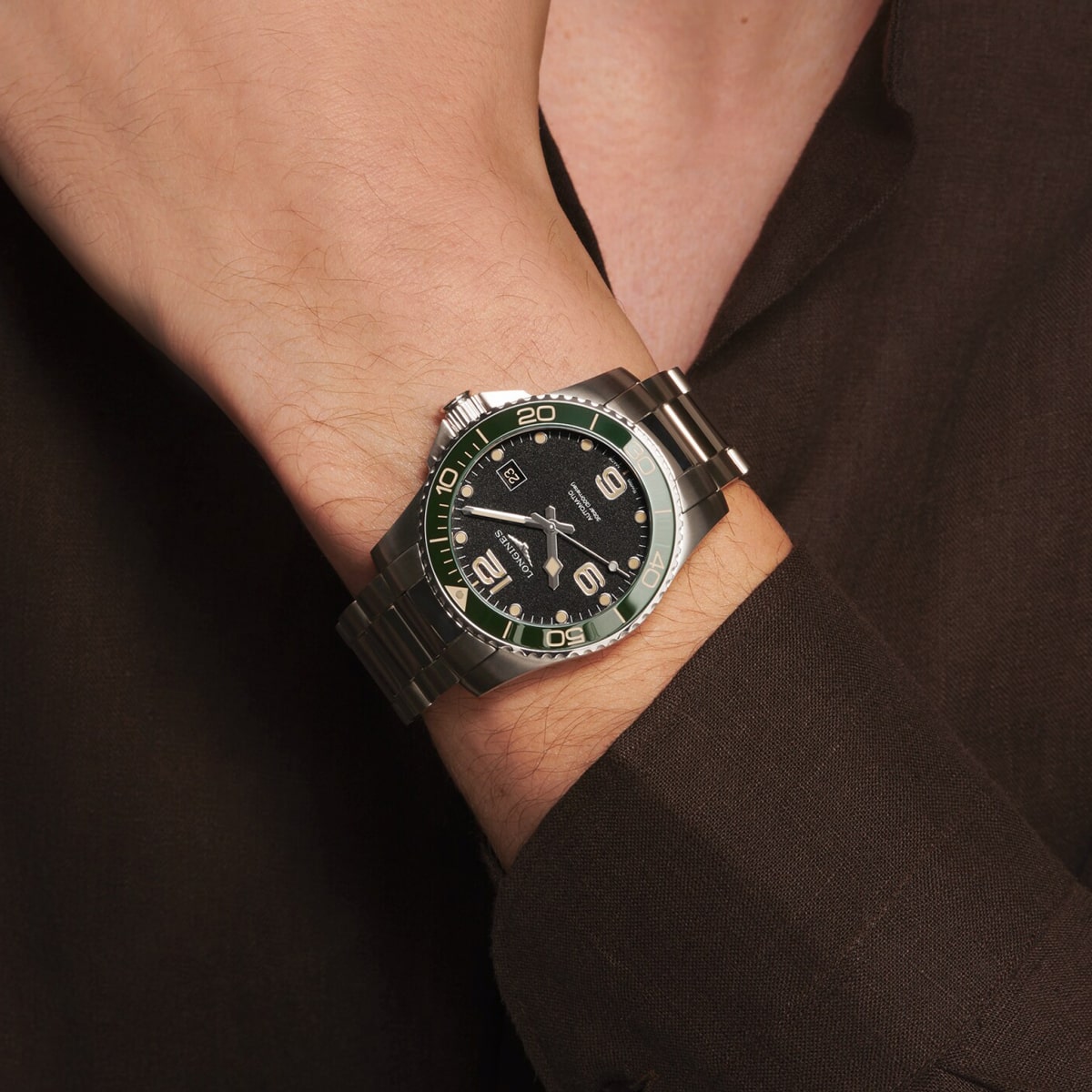 Eine Person, die ein dunkles Hemd trägt, präsentiert eine silberne HydroConquest-Armbanduhr mit grüner und schwarzer Lünette und schwarzem Zifferblatt, die die Uhrzeit am linken Handgelenk anzeigt.