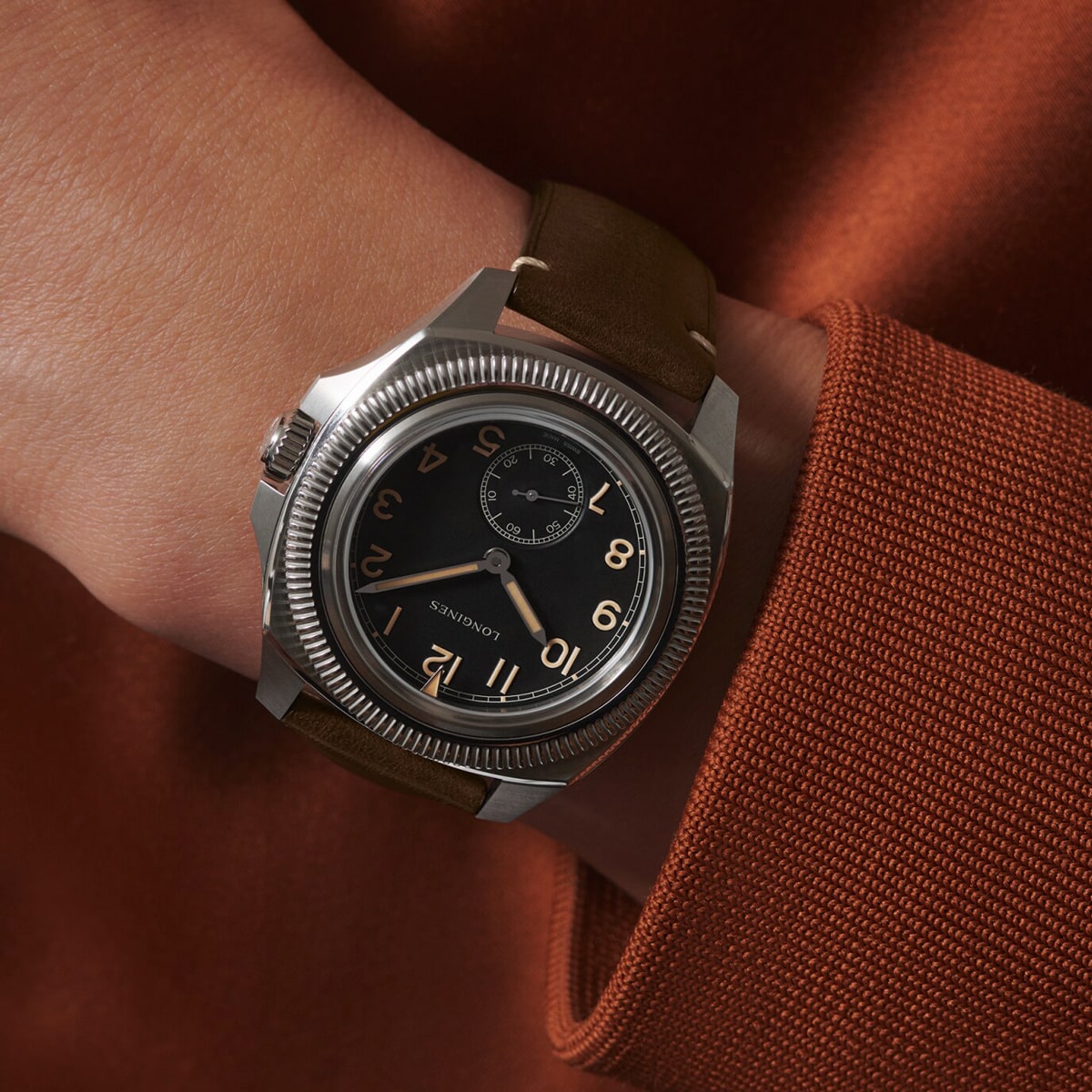 2025 © LONGINES Eine Person, die eine Pilot-Majetek-Uhr mit einem braunen Lederarmband und einem schwarzen Zifferblatt mit arabischen Ziffern trägt, bekleidet mit einem orangefarbenen langärmeligen Kleidungsstück.