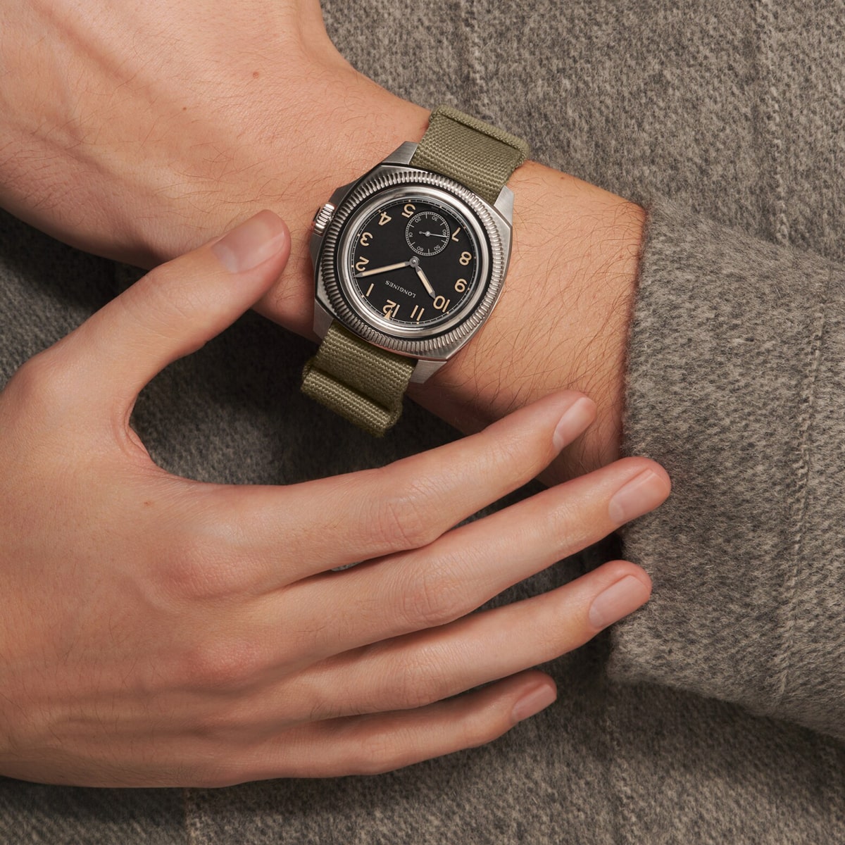 2025 © LONGINES Eine Person, die ein graues, langärmeliges Hemd trägt, zeigt ein Handgelenk mit einer Pilot Majetek, die ein rundes, schwarzes Zifferblatt und ein olivgrünes Armband hat, während ihre andere Hand sanft das Handgelenk mit der Uhr berührt.