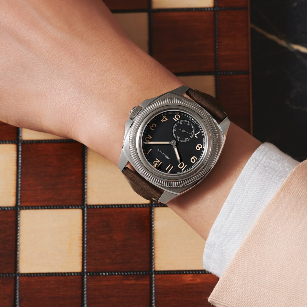 2025 © LONGINES Eine Person, die eine Pilot Majetek-Armbanduhr mit braunem Lederarmband und schwarzem Zifferblatt mit großen arabischen Ziffern trägt, steht über einem hölzernen Schachbrett.